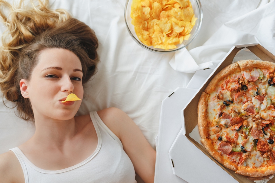 Girl eating chips on the bed, standing next to the pizza. Girl looking at camera lying on white bed
